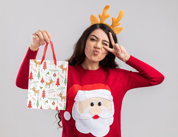 Pewna siebie młoda ładna dziewczyna ubrana w opaskę z poroża renifera i sweter świętego mikołaja trzyma torbę z prezentami bożonarodzeniowymi, patrząc na symbol znaku v w pobliżu mrugającego okiem