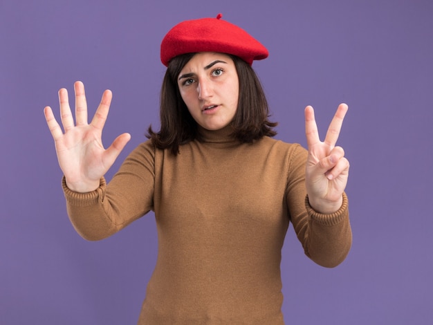 Pewna siebie młoda ładna kaukaska dziewczyna w kapeluszu beret gestykuluje siedem palcami na fioletowo