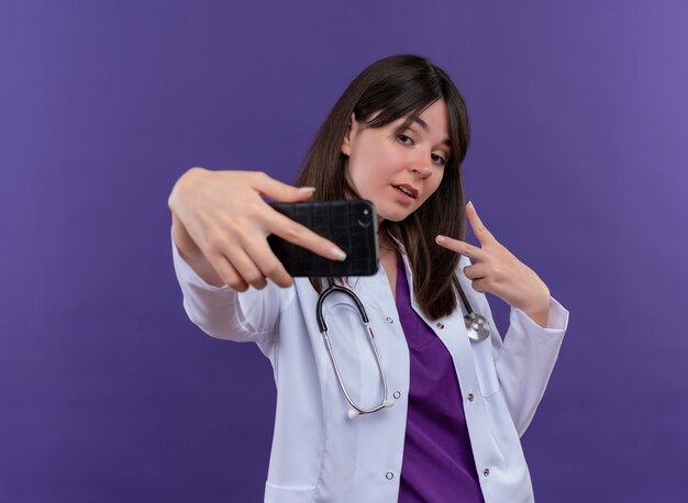 Pewna siebie młoda lekarka w szacie medycznej ze stetoskopem patrzy na telefon i gestykuluje znak zwycięstwa palcami na izolowanym fioletowym tle z miejsca na kopię