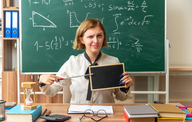 Pewna siebie młoda nauczycielka siedzi przy stole z narzędziami szkolnymi wskazuje na mini tablicę z kijem wskaźnikowym w klasie