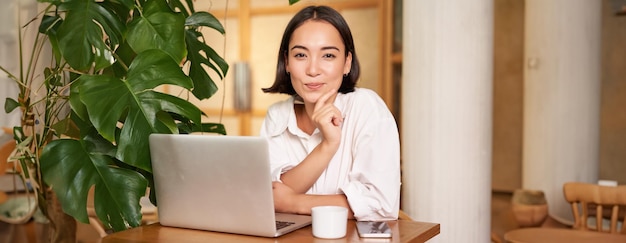Pewna siebie młoda stylowa kobieta z laptopem siedząca w kawiarni i pracująca jako freelancer w przestrzeni coworkingowej