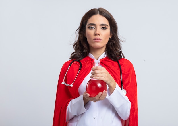 Pewna siebie superkobieta w mundurze lekarza z czerwoną peleryną i stetoskopem trzymająca czerwony płyn chemiczny w szklanej kolbie na białej ścianie