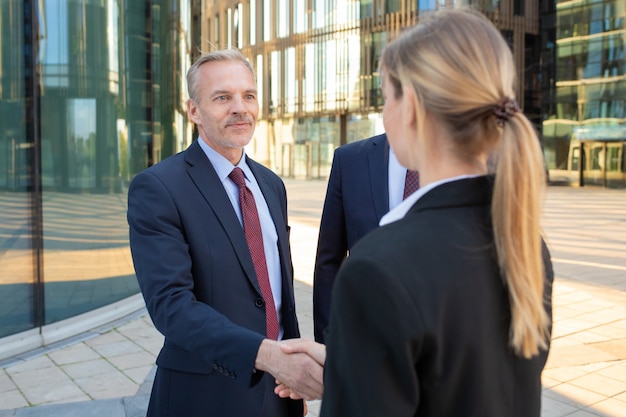 Pewni siebie partnerzy biznesowi stojący w pobliżu biurowców, ściskający ręce, spotykający się i rozmawiający w mieście. Omówienie umowy i koncepcja partnerstwa