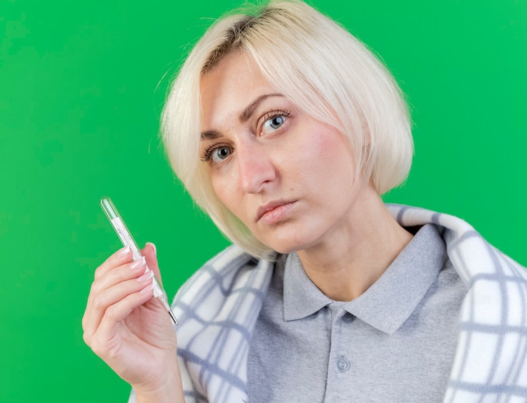 Bezpłatny plik Zdjęcie pewnie młoda blondynka chora kobieta owinięta w kratę trzymając termometr patrząc z przodu na białym tle na zielonej ścianie