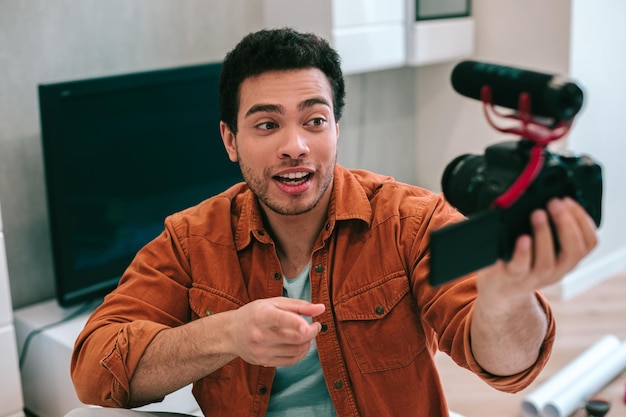 Pewny siebie, atrakcyjny młody mężczyzna w koszuli i t-shircie wskazujący na kamerę podczas nagrywania wideo ze sobą