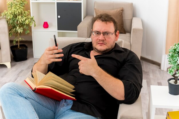 Pewny siebie dorosły słowiański mężczyzna w okularach optycznych siedzi na fotelu trzymając książkę na nogach i wskazując na telefon w salonie