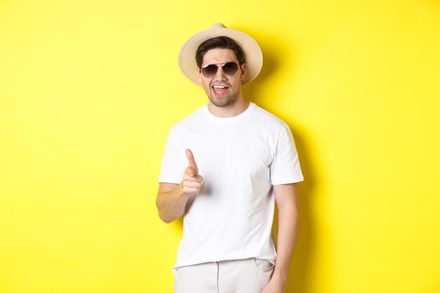 Pewny siebie i bezczelny facet na wakacjach flirtuje z tobą, wskazując palcem na aparat i mrugając, ubrany w letni kapelusz z okularami przeciwsłonecznymi, żółte tło
