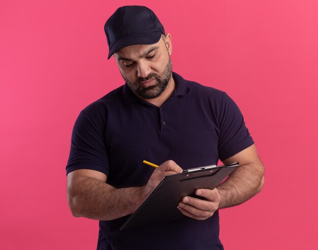 Pewny siebie mężczyzna w średnim wieku w mundurze i czapce piszący coś w schowku na różowej ścianie