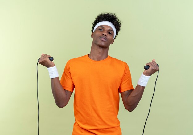 Pewny siebie młody afro-amerykański sportowy mężczyzna noszenie opaski i opaski na rękę trzymając skakankę na białym tle na zielonym tle