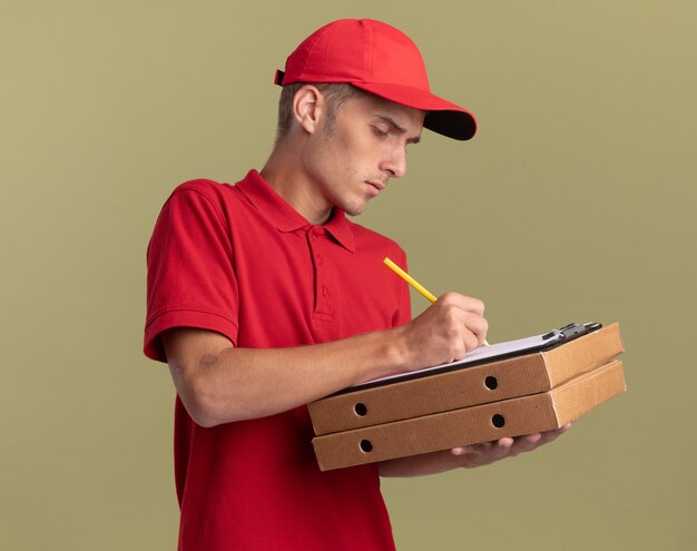Pewny siebie młody blond chłopiec dostawy pisze w schowku ołówkiem trzymając pudełka po pizzy na oliwkowej zieleni