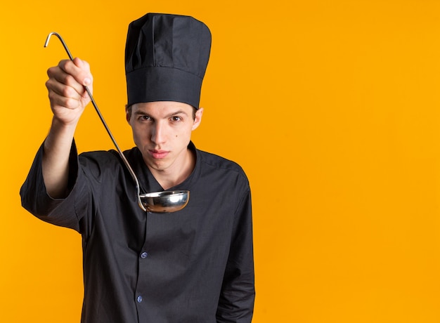 Pewny siebie młody blond mężczyzna kucharz w mundurze szefa kuchni i czapce, patrząc na kamerę wyciągającą kadzi w kierunku kamery izolowanej na pomarańczowej ścianie z miejscem na kopię