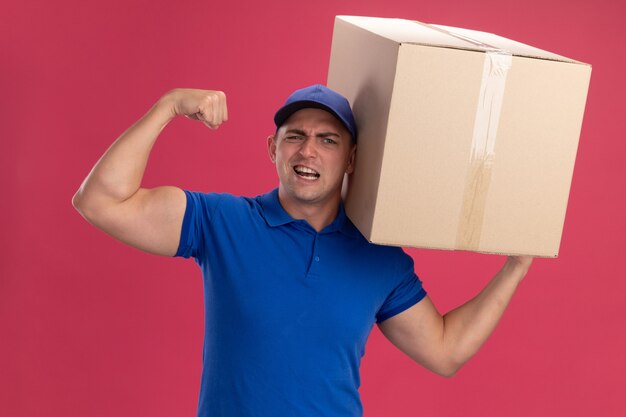 Pewny siebie młody dostawca ubrany w mundur z czapką trzyma duże pudełko pokazujące silny gest na różowej ścianie
