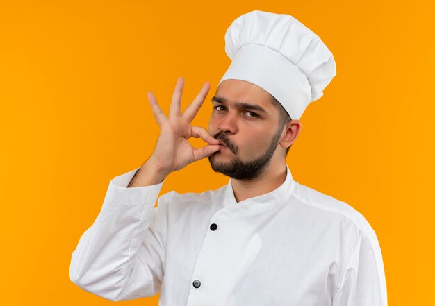Pewny siebie młody kucharz w mundurze szefa kuchni robi smaczny gest na pomarańczowej ścianie