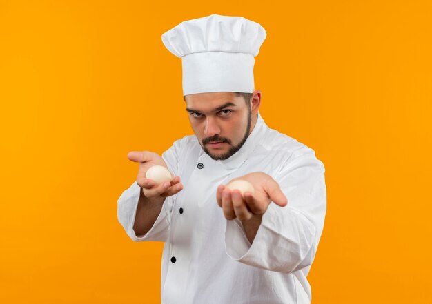 Pewny siebie młody kucharz w mundurze szefa kuchni rozciągający jajka w kierunku izolowanej na pomarańczowej ścianie z miejscem na kopię