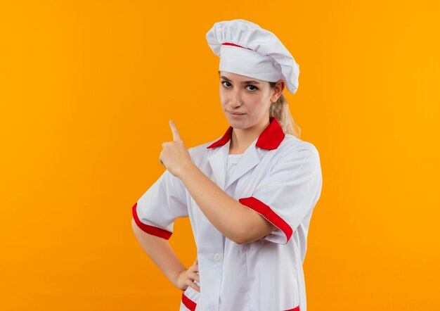 Pewny siebie młody ładny kucharz w mundurze szefa kuchni, kładąc rękę na talii, skierowaną w górę na białym tle na pomarańczowej ścianie