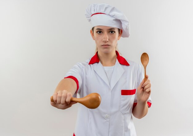 Pewny siebie młody ładny kucharz w mundurze szefa kuchni trzymający i wyciągający łyżki izolowane na białej ścianie