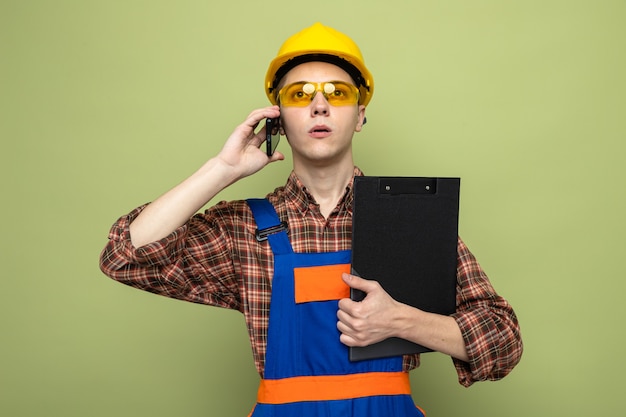 Bezpłatne zdjęcie pewny siebie młody mężczyzna budowniczy trzymający schowek w mundurze w okularach mówi przez telefon