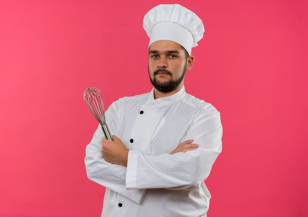 Pewny siebie młody mężczyzna kucharz w mundurze szefa kuchni, stojący z zamkniętą postawą i trzymający trzepaczkę odizolowaną na różowej ścianie