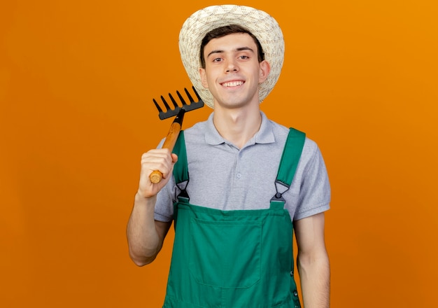 Pewny siebie młody ogrodnik mężczyzna w kapeluszu ogrodniczym trzyma grabie na ramieniu