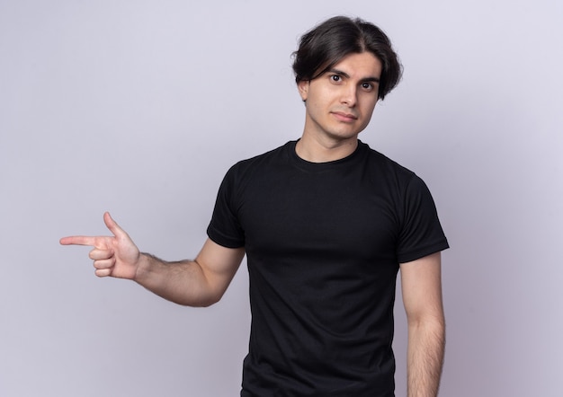 Pewny siebie młody przystojny facet ubrany w czarny t-shirt wskazuje z boku na białym tle na białej ścianie z miejscem na kopię