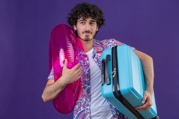 Pewny siebie młody przystojny kręcone podróżnik mężczyzna trzyma walizkę i pierścień do pływania patrząc na lewą stronę na odizolowanej fioletowej przestrzeni z kopią miejsca