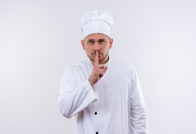 Pewny siebie młody przystojny kucharz w mundurze szefa kuchni gestykuluje ciszę na białej ścianie z miejscem na kopię