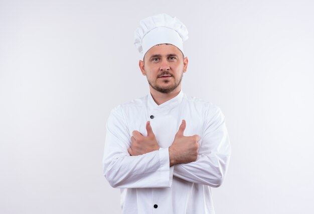 Pewny siebie młody przystojny kucharz w mundurze szefa kuchni stojący z zamkniętą postawą pokazując kciuk do góry na białym tle na białej ścianie