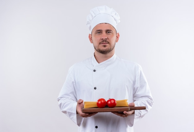 Bezpłatne zdjęcie pewny siebie młody przystojny kucharz w mundurze szefa kuchni trzymający deskę do krojenia z pomidorami i makaronem spaghetti na białym tle na białej ścianie