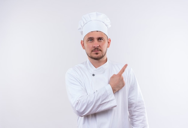 Pewny siebie młody przystojny kucharz w mundurze szefa kuchni, wskazując na bok na białym tle na białej ścianie