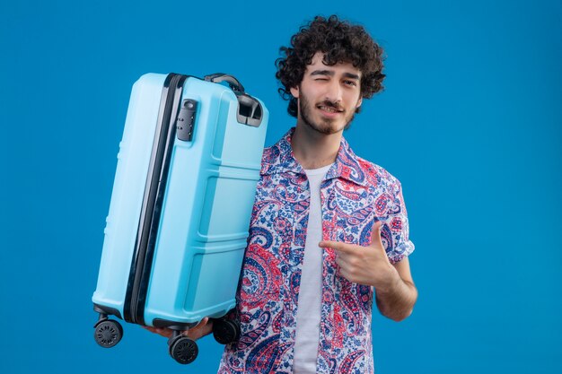Pewny siebie młody przystojny podróżnik mężczyzna trzyma walizkę, wskazując na to i mrugając na odizolowanej niebieskiej przestrzeni z kopią miejsca