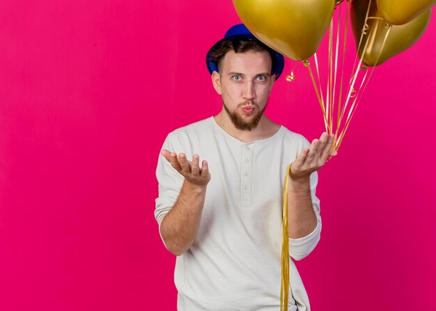 Pewny siebie młody przystojny słowiański imprezowicz w kapeluszu imprezowym, trzymając balony patrząc z przodu, pokazując pustą dłoń robi gest pocałunku na różowej ścianie z miejsca na kopię