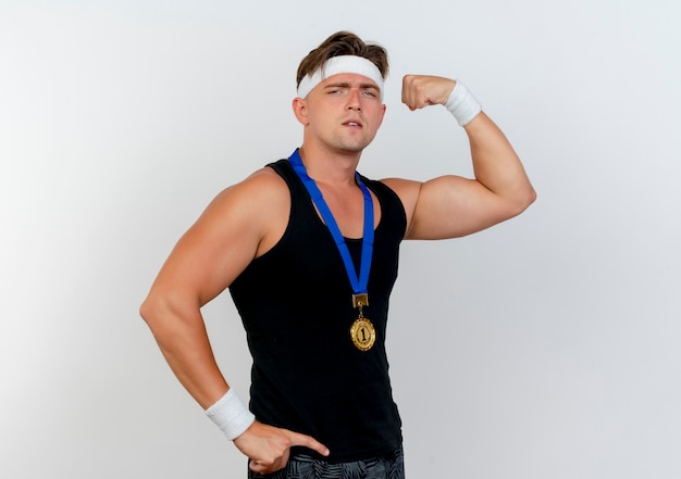 Pewny siebie młody przystojny sportowy mężczyzna noszący opaskę i opaski na nadgarstek oraz medal na szyi, kładący rękę na talii i gestykulujący silny na białym tle