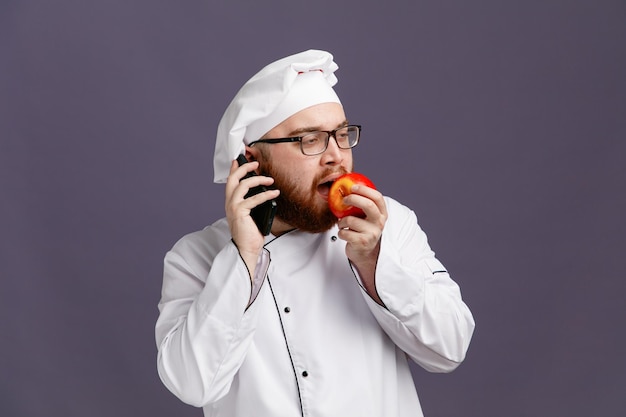 Bezpłatne zdjęcie pewny siebie młody szef kuchni w mundurze okularowym i czapce, patrzący na bok, jedzący jabłko podczas rozmowy przez telefon na fioletowym tle