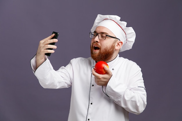 Pewny siebie młody szef kuchni w mundurze okularowym i czapce trzymającej jabłko i telefon komórkowy, biorąc selfie na białym tle na fioletowym tle