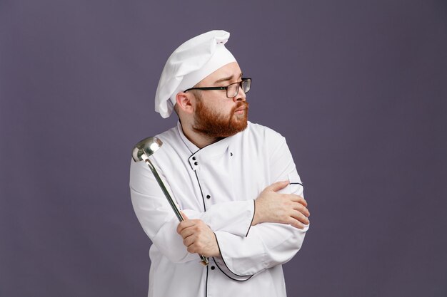 Pewny siebie młody szef kuchni w okularach mundurowych i czapce, patrząc na bok trzymając kadzi, trzymając rękę na ramieniu na fioletowym tle