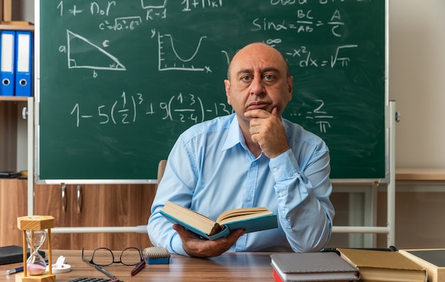 Bezpłatne zdjęcie pewny siebie nauczyciel w średnim wieku siedzi przy stole z przyborami szkolnymi, trzymając książkę chwycił podbródek w klasie