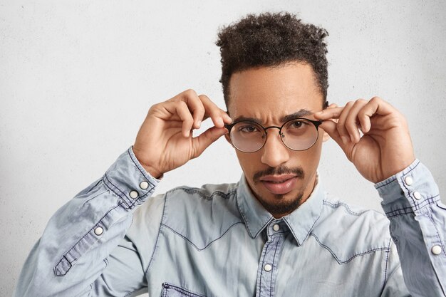 Pewny siebie przedsiębiorca rasy mieszanej trzyma ręce na oprawkach okularów, przygląda się uważnie