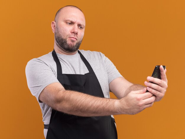 Pewny siebie słowiański fryzjer w średnim wieku w mundurze robi selfie na pomarańczowej ścianie