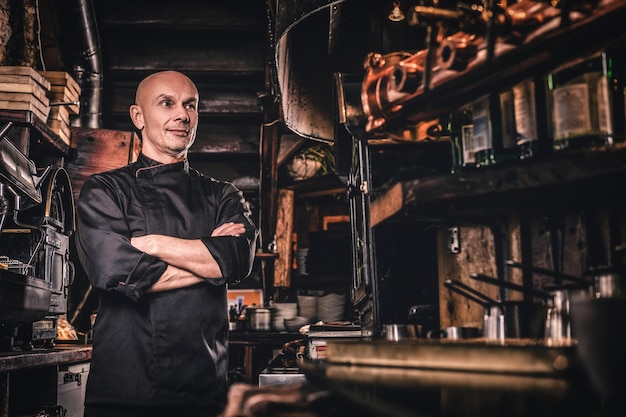 Pewny siebie szef kuchni w mundurze pozuje ze skrzyżowanymi rękami i odwraca wzrok w kuchni restauracji.