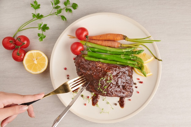 Pieczone, grillowane żeberka wieprzowe z letniego grilla podawane z warzywami, szparagami, młodą marchewką, świeżymi pomidorami, przyprawami na białym talerzu. kobieta ręce z widelcem i nożem, jedzenie żeber. widok z góry.