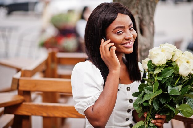 Piękna afroamerykanka trzyma bukiet kwiatów białych róż na randki w mieście Czarna kobieta z bukietem kwiatów z telefonem komórkowym
