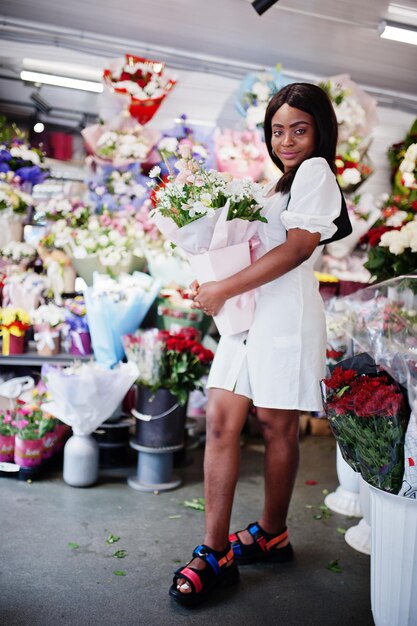 Piękna afroamerykańska dziewczyna w delikatnej białej sukni z bukietem kwiatów w dłoniach stojąca na tle kwiatów w kwiaciarniCzarna kwiaciarnia żeńska