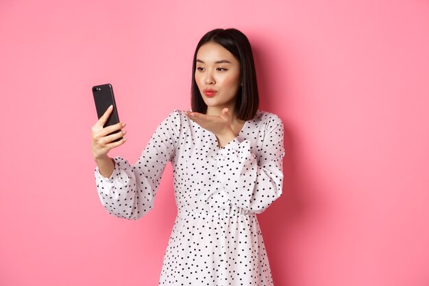 Piękna azjatycka dziewczyna korzystająca z aplikacji filtrów fotograficznych i robiąca selfie na smartfonie, pozująca w uroczej sukience na różowym tle