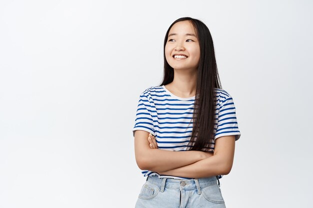 Piękna azjatycka dziewczyna patrząca na bok ze szczęśliwym uśmiechem, skrzyżowanymi ramionami na piersi, patrząca na coś po lewej stronie, stojąca na białym tle