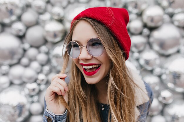 Piękna biała dziewczyna śmiejąc się i bawiąc się jej blond włosami na błyszczącej ścianie. Zdjęcie słodkie modelki w modnym czerwonym kapeluszu, wyrażające szczęśliwe emocje.