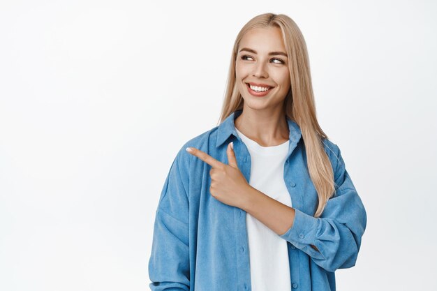 Piękna blond kobieta uśmiecha się palcem wskazującym i patrzy w lewo na logo firmy pokazujące reklamę stojącą na białym tle