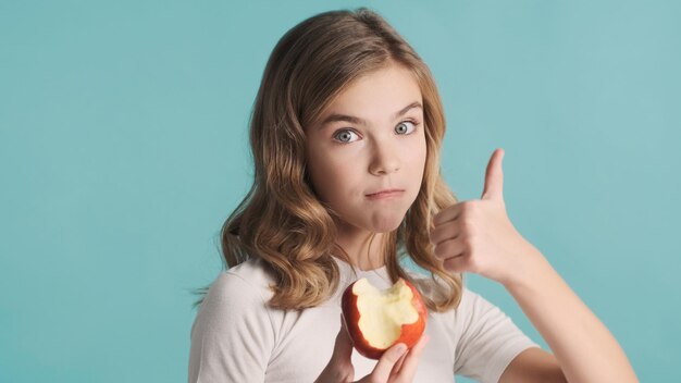 Piękna blond nastolatka dziewczyna z falowanymi włosami, jedzenie pyszne jabłko, trzymając kciuk na niebieskim tle. Lubię to