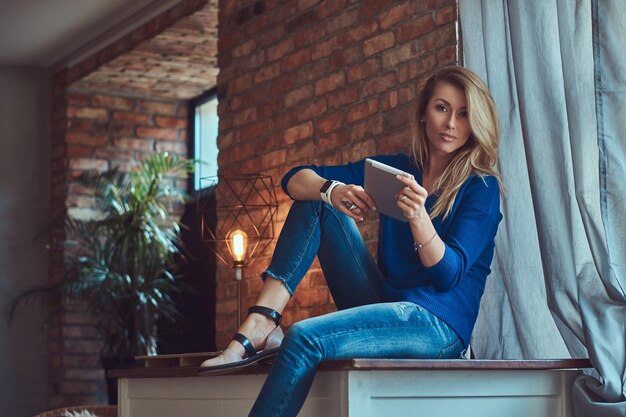 Piękna blondynka blogerka trzyma tablet, siedząc na stole przy ścianie z cegły w studio z wnętrzem na poddaszu.