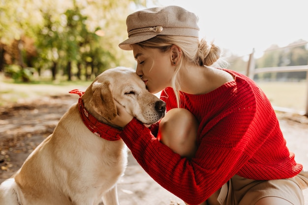 Piękna blondynka całuje swojego uroczego psa w jesiennym słonecznym parku. Stylowa młoda kobieta w czerwonym swetrze i modnym kapeluszu czule trzymając zwierzaka.