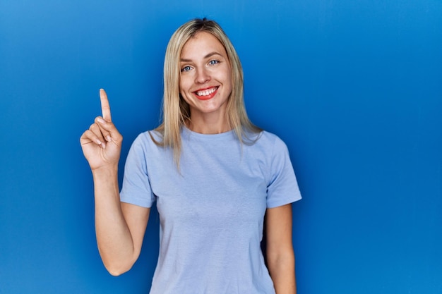 Bezpłatne zdjęcie piękna blondynka ubrana w swobodną koszulkę na niebieskim tle, pokazująca i wskazująca palcem numer jeden, uśmiechając się pewna siebie i szczęśliwa.